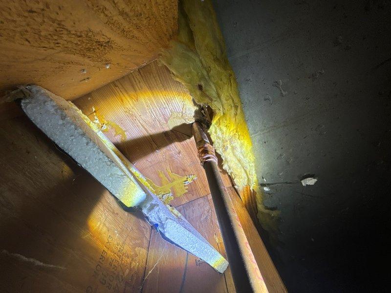 Responded to service call for exterior hose bibb that was difficult to operate and experiencing leakage issues when shutting off. Accessed the outdoor spigot from interior crawl space to diagnose the problem. Upon inspection, found the supply line and shutoff valve assembly in the basement area beneath the exterior wall penetration. The copper supply piping and associated connections showed signs of age and wear. Evaluated the valve mechanism and determined the root cause of the operational difficulty and leaking condition. Documented the existing installation with photographic evidence showing the supply line routing through floor joists with surrounding insulation, as well as the pipe penetration through the foundation wall. The images captured the current state of the plumbing assembly, insulation placement, and structural elements for reference. Provided assessment of necessary repairs to restore proper function and eliminate the leak when closing the valve.