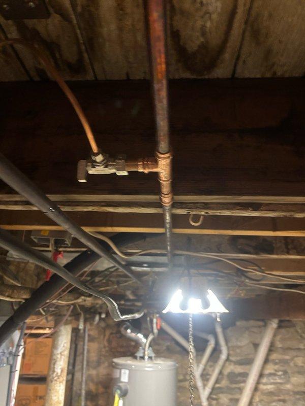 Responded to emergency call for active water leak in basement of century-old home. Upon arrival, found hot water line had been accidentally pierced during recent AC humidifier installation, causing water to spray throughout the basement area. Homeowner had temporarily stopped the leak by shutting off hot water supply at the water heater. Inspected the damaged copper supply line running through the basement ceiling joists and identified the puncture point where the line had been compromised during the previous week's HVAC work. Assessed the surrounding plumbing infrastructure, including multiple copper pipes and connections in the unfinished basement space. Repaired the pierced hot water line by cutting out the damaged section and installing new copper piping with properly soldered joints and fittings. Restored water supply and pressure tested all connections to ensure no additional leaks were present. Verified proper operation of the hot water system and confirmed the humidifier supply