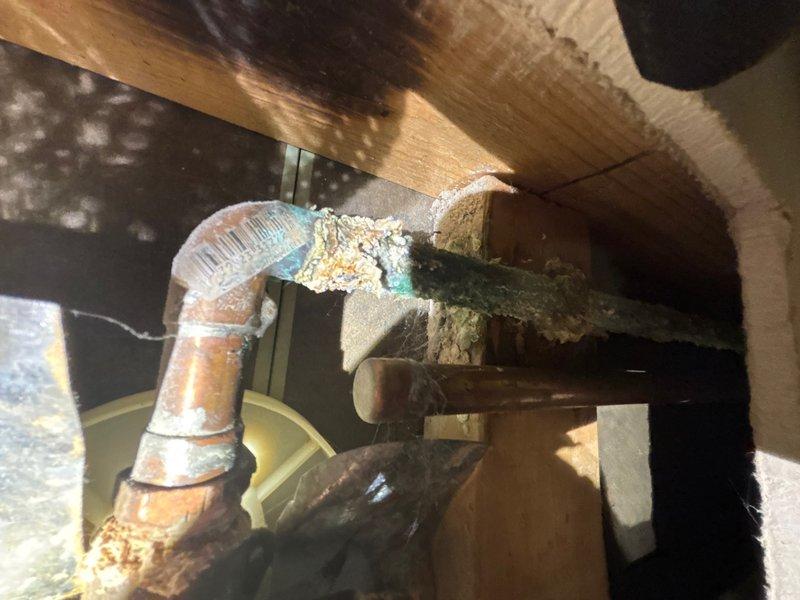 Responded to service call for reported leak under kitchen sink drain. Upon arrival, inspected kitchen sink drain assembly and surrounding cabinetry for active leaks and water damage. Examined drain connections, trap assembly, and supply lines for signs of moisture or failure.

Investigation revealed water intrusion issues extending beyond the initially reported kitchen sink location. Inspection of crawl space or subfloor area disclosed significant moisture concerns affecting structural components. Observed dark staining and discoloration on wooden floor joists and beams indicating prolonged water exposure. Located copper supply piping with extensive condensation buildup, evidenced by heavy frost or mineral deposit accumulation on pipe surfaces and elbow joints.

Documented conditions with photographic evidence showing moisture damage to flooring transition areas, with visible water staining on carpeted surfaces near doorway thresholds. Additional documentation captured extent of pipe c