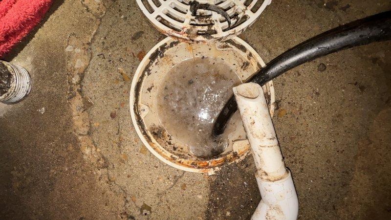 Responded to clogged basement floor drain. Upon arrival, found standing water in white PVC drain assembly with visible sediment buildup and brown staining around drain body and interior surfaces. Cleared blockage from drain line, restoring proper water flow and drainage function.