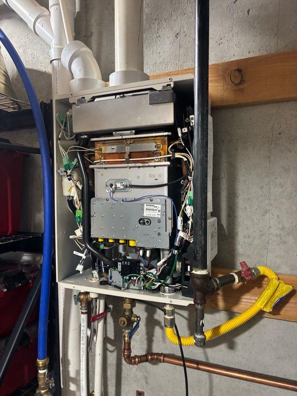 Responded to service call for tankless water heater replacement evaluation in basement location. Upon arrival, inspected existing wall-mounted tankless unit with front access panel removed to assess internal components including copper heating elements, control board, electrical connections, and brass fittings. Documented installation configuration including white PVC venting system, yellow corrugated gas line, and copper water supply connections mounted on concrete wall with wooden support beam. Evaluated unit condition and provided estimate for tankless water heater replacement based on customer requirements and existing gas utility infrastructure.