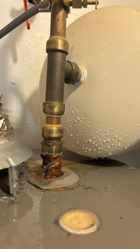 Responded to reported water leakage behind tanked water heater in basement with water accumulation on laminated flooring. Upon inspection, identified significant rust and corrosion at the pipe connection point at the base of the tank where the vertical supply line connects, with visible water droplets on tank exterior and pooling around the corroded connection. The deteriorated fitting showed orange-brown discoloration indicating advanced corrosion compromising the integrity of the connection. Documented the condition of the plumbing assembly including brass compression fittings, flexible braided supply lines, shut-off valve, and mounting hardware for assessment and repair recommendations.
