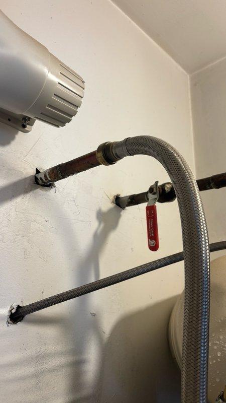 Responded to reported water leakage behind tanked water heater in basement with water accumulation on laminated flooring. Upon inspection, identified significant rust and corrosion at the pipe connection point at the base of the tank where the vertical supply line connects, with visible water droplets on tank exterior and pooling around the corroded connection. The deteriorated fitting showed orange-brown discoloration indicating advanced corrosion compromising the integrity of the connection. Documented the condition of the plumbing assembly including brass compression fittings, flexible braided supply lines, shut-off valve, and mounting hardware for assessment and repair recommendations.
