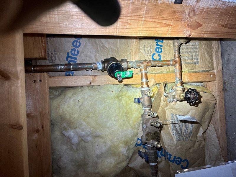 Responded to water leak complaint originating from bottom of water heater tank located in basement utility area. Upon inspection, confirmed active leak from lower tank section of residential water heater installed in 2021. Assessed plumbing connections, supply lines, and shut-off valves in wall cavity featuring copper piping with brass fittings and pressure regulation components. Documented existing conditions including water heater installation with associated plumbing infrastructure, copper supply lines with braided stainless steel flex connectors, and mechanical room layout. Photographed plumbing assembly and water heater unit to record leak source and equipment configuration for service documentation purposes.