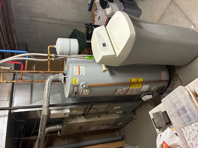 Responded to water leak complaint originating from bottom of water heater tank located in basement utility area. Upon inspection, confirmed active leak from lower tank section of residential water heater installed in 2021. Assessed plumbing connections, supply lines, and shut-off valves in wall cavity featuring copper piping with brass fittings and pressure regulation components. Documented existing conditions including water heater installation with associated plumbing infrastructure, copper supply lines with braided stainless steel flex connectors, and mechanical room layout. Photographed plumbing assembly and water heater unit to record leak source and equipment configuration for service documentation purposes.