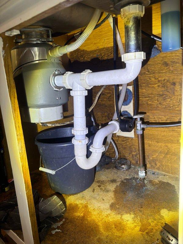 Responded to service call for garbage disposal leak and clogged drainage system affecting kitchen and bathroom fixtures. Upon inspection, identified active leak from garbage disposal unit with visible yellow discharge into containment bucket. Assessed P-trap configuration and disposal mounting assembly for integrity issues. Addressed clogged drainage in bathroom sink and kitchen plumbing system. Cleared blockages from waste lines, inspected disposal unit connections and mounting flange for proper seal, and verified P-trap assembly for debris accumulation. Documented existing water damage on cabinet floor indicating previous leakage issues. Tested drainage flow through garbage disposal and connected waste lines to ensure proper function and eliminate backup conditions.