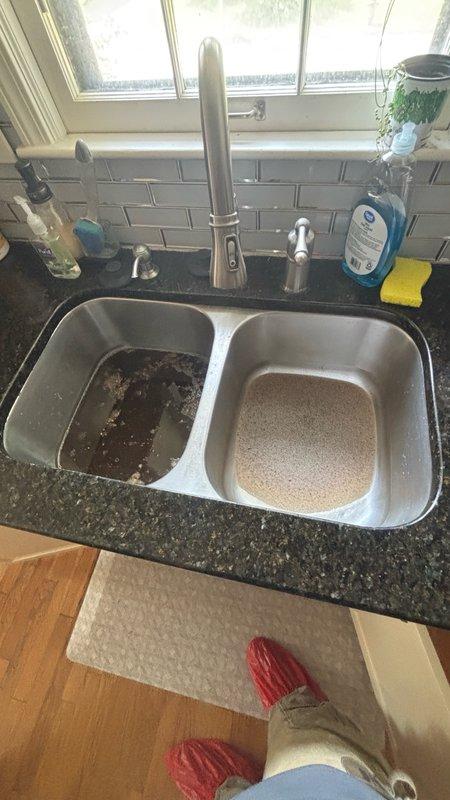 Responded to kitchen sink backup caused by clogged drain line. Upon arrival, found left basin retaining standing water with visible food debris and dark residue accumulation. Accessed cabinet area beneath sink to inspect drain assembly and P-trap configuration. Observed moisture damage and discoloration on cabinet interior materials indicating previous or ongoing drainage issues. Cleared blockage from drain line, removed food particle buildup, and restored proper water flow through both basins. Verified drainage function and recommended monitoring cabinet condition for potential water damage repairs.