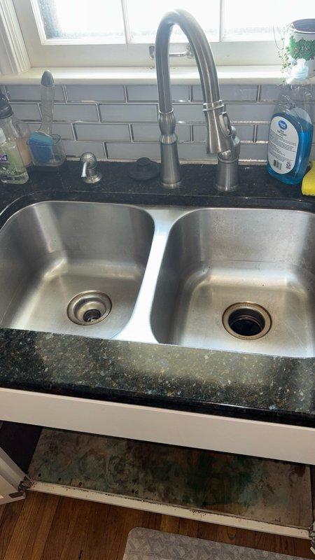 Responded to kitchen sink backup caused by clogged drain line. Upon arrival, found left basin retaining standing water with visible food debris and dark residue accumulation. Accessed cabinet area beneath sink to inspect drain assembly and P-trap configuration. Observed moisture damage and discoloration on cabinet interior materials indicating previous or ongoing drainage issues. Cleared blockage from drain line, removed food particle buildup, and restored proper water flow through both basins. Verified drainage function and recommended monitoring cabinet condition for potential water damage repairs.