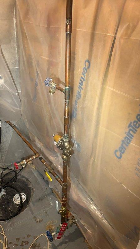 Completed plumbing maintenance visit involving inspection and documentation of copper piping installation in basement utility area. System includes vertical copper supply lines with multiple brass ball valves (blue and red handled), T-fittings, and tagged valve components installed against foam insulation board with vapor barrier. Circulator pump or similar equipment present at floor level with electrical connections. All components photographed for record, piping assembly and valve configuration verified operational.
