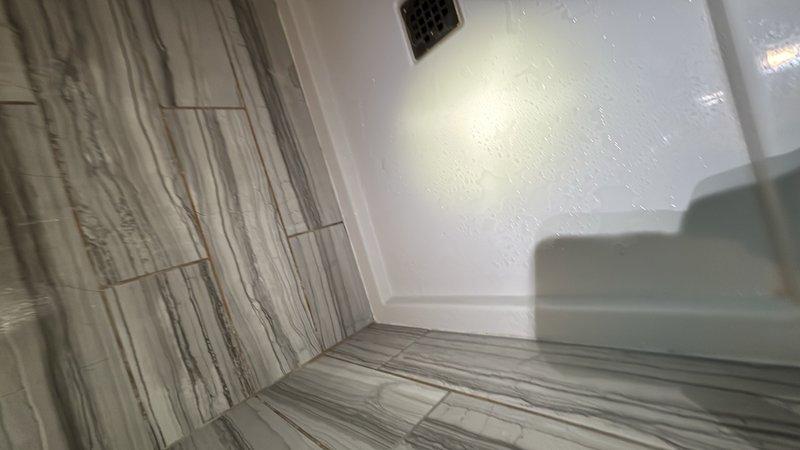 Responded to report of water leaking from under shower pan after use. Performed thorough inspection of shower enclosure, including examination of tile work, grout lines, and shower pan integrity. Inspected modern marble-pattern porcelain tile installation on walls and floor, checking for compromised grout joints and potential water intrusion points. Examined glass shower enclosure seals and mounting hardware for proper installation and waterproofing. Documented findings with photos showing tile condition and enclosure assembly. Identified potential leak source and provided recommendations for repair to prevent continued water migration beneath shower pan.