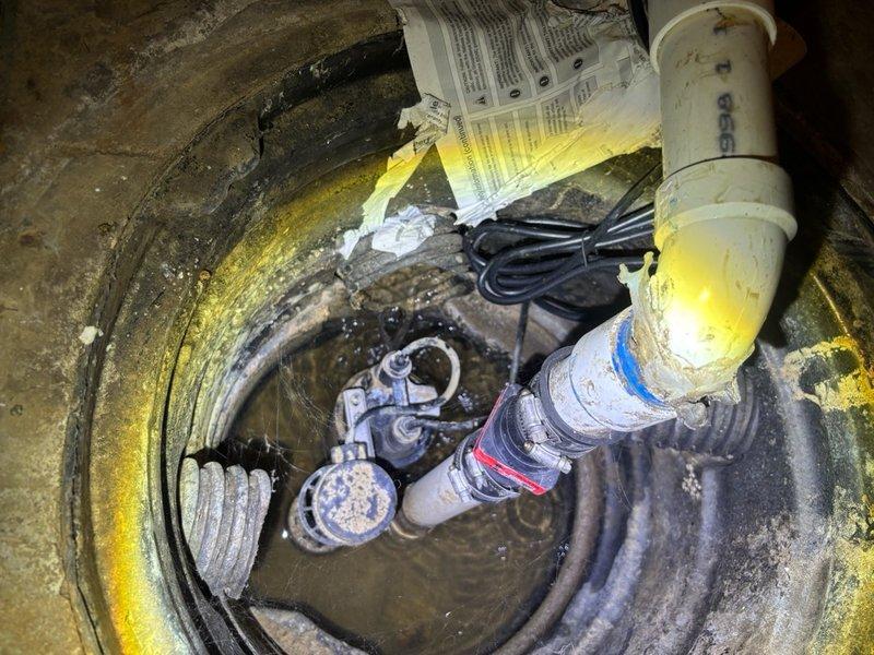 Responded to service call for sump pump activating but water pooling adjacent to unit in sub basement. Upon inspection, found existing sump pump system with visible mineral deposits and sediment accumulation throughout pit. Pump was cycling on as reported, but discharge system required evaluation due to water backup. Assessed PVC discharge piping, pump float mechanism, and check valve operation. Pit showed signs of age with heavy buildup on walls and standing water at base. Documented existing conditions including pump assembly, electrical connections, and discharge plumbing configuration for repair recommendations.