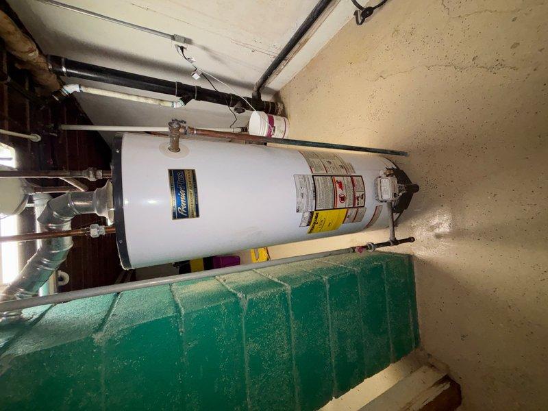 Conducted on-site assessment for water heater replacement quote. Inspected existing white cylindrical water heater unit located in basement utility room, currently operational but requiring replacement evaluation. Documented unit specifications, manufacturer information, and existing plumbing configuration including copper supply lines, pressure relief valve, and top-mounted connections. Identified significant corrosion and rust deterioration on copper piping system, particularly at pipe joints and connections passing through floor penetrations. Photographed water heater installation, surrounding HVAC ductwork, and corroded plumbing components to support replacement quotation. Evaluated utility room access, clearances, and existing infrastructure to determine replacement requirements and specifications.