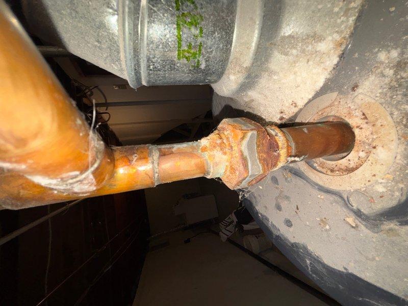 Conducted on-site assessment for water heater replacement quote. Inspected existing white cylindrical water heater unit located in basement utility room, currently operational but requiring replacement evaluation. Documented unit specifications, manufacturer information, and existing plumbing configuration including copper supply lines, pressure relief valve, and top-mounted connections. Identified significant corrosion and rust deterioration on copper piping system, particularly at pipe joints and connections passing through floor penetrations. Photographed water heater installation, surrounding HVAC ductwork, and corroded plumbing components to support replacement quotation. Evaluated utility room access, clearances, and existing infrastructure to determine replacement requirements and specifications.