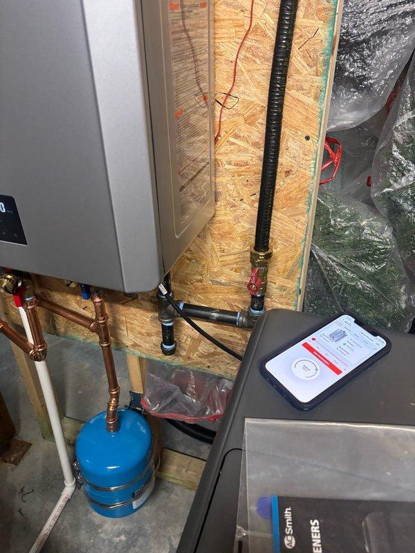 Completed installation of Rinnai RXP199 tankless water heater with RWM200 wireless module in unfinished mechanical space. Installation included mounting unit to OSB wall, running copper supply lines with brass fittings, installing blue expansion tank at floor level, and routing black corrugated flexible venting secured with zip ties. Configured PVC condensate drainage system through ceiling joists with proper support brackets and integrated wireless monitoring module for remote system management.