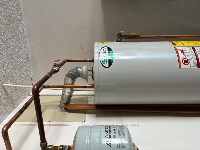 Responded to service call for gas water heater making beeping noise in garage. Upon arrival, inspected the gas-fired water heater installation and associated components. Evaluated the unit's operational status, checked control board functions, and verified proper gas supply and venting. Examined the expansion tank, copper supply lines, and all plumbing connections for proper installation and function. Tested the water heater's electronic controls and safety features to identify the source of the audible alarm. Photographed the installation showing the horizontal tank configuration with copper piping, galvanized connections, and the Amtrol expansion tank mounted below on the platform. Verified all components were functioning within normal parameters and addressed the beeping concern.