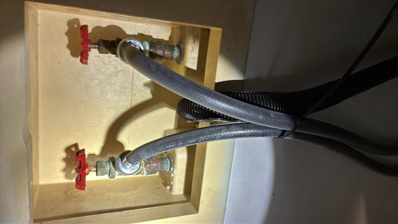 Completed comprehensive plumbing inspection of residential water supply system and water treatment equipment. Inspected wall-mounted washing machine supply connection box with dual shut-off valves and flexible supply lines, verifying proper operation of red-handled valves and integrity of braided and corrugated hose connections. Evaluated water softener system with digital control panel displaying current water flow reading of 43.4 gallons, confirmed operational status of control buttons and salt level monitoring system. All plumbing fixtures and connections found to be functioning within normal parameters with no immediate repairs required.