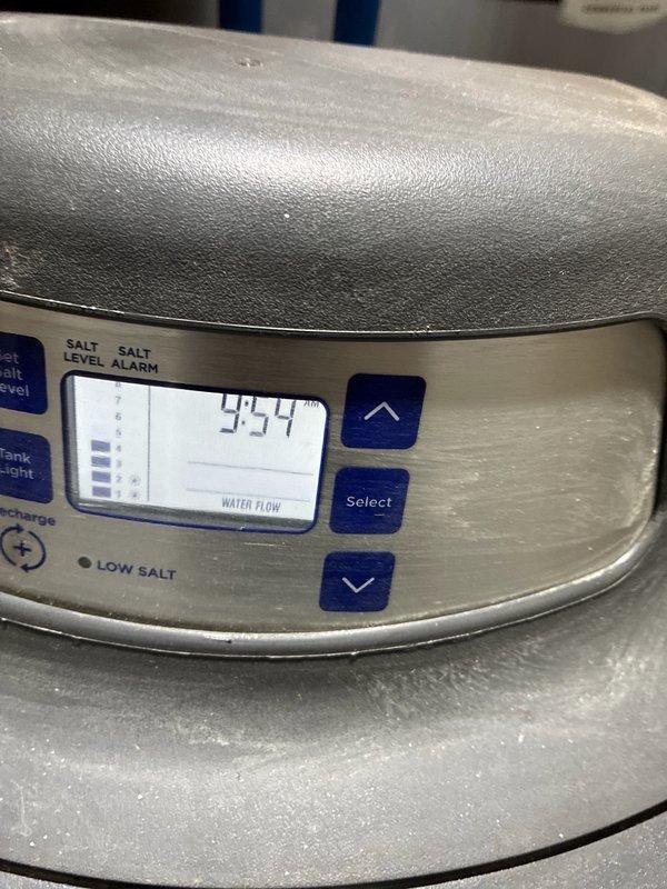 Completed comprehensive plumbing inspection of residential water supply system and water treatment equipment. Inspected wall-mounted washing machine supply connection box with dual shut-off valves and flexible supply lines, verifying proper operation of red-handled valves and integrity of braided and corrugated hose connections. Evaluated water softener system with digital control panel displaying current water flow reading of 43.4 gallons, confirmed operational status of control buttons and salt level monitoring system. All plumbing fixtures and connections found to be functioning within normal parameters with no immediate repairs required.