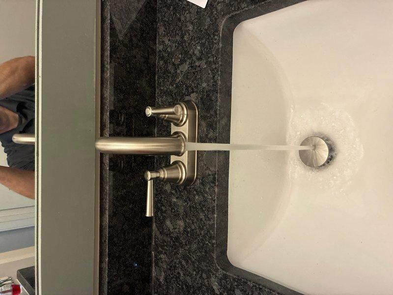 Completed installation of customer-supplied faucet assembly with pop-up drain mechanism in bathroom vanity. The wall-mounted faucet features a brushed nickel finish with dual lever handles and horizontal spout, installed above a white rectangular vessel sink against dark granite backsplash. All connections were secured, tested for proper water flow and drainage, and verified leak-free operation upon completion.