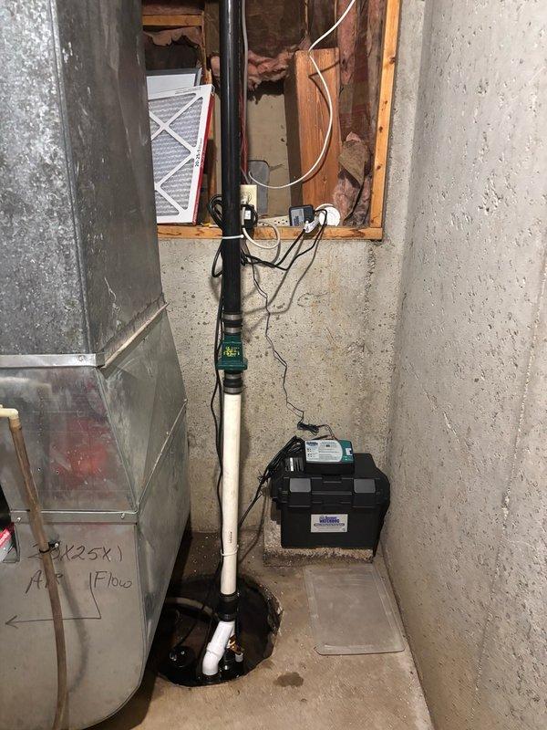 Installed Watchdog sump pump system with customer-supplied backup battery in basement utility area. System includes white PVC discharge piping mounted vertically along concrete wall with proper mounting brackets, black cylindrical sump basin positioned at floor level, and complete electrical connections for primary pump and battery backup operation. All wiring secured and routed through wall opening to power source, ensuring reliable operation during power outages and normal conditions.
