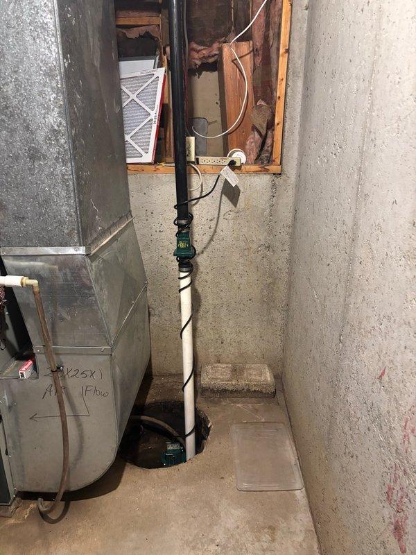 Installed Watchdog sump pump system with customer-supplied backup battery in basement utility area. System includes white PVC discharge piping mounted vertically along concrete wall with proper mounting brackets, black cylindrical sump basin positioned at floor level, and complete electrical connections for primary pump and battery backup operation. All wiring secured and routed through wall opening to power source, ensuring reliable operation during power outages and normal conditions.