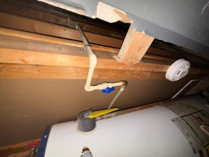 Responded to service call for water heater replacement estimate in attic/crawl space location. Upon arrival, accessed confined utility area and assessed existing water heater installation. Documented current plumbing configuration including copper supply lines with brass fittings, red-handled shut-off valve, white PVC venting system with elbow joints and blue connectors, and metal strapping securing the unit. Observed moisture present on vertical PVC piping and noted opening in ceiling material near installation area. Evaluated structural clearances, wooden joist spacing, and access requirements for 40-gallon capacity replacement unit. Photographed existing installation setup, pipe routing, valve locations, and venting configuration to prepare accurate replacement estimate. Provided customer with assessment findings and replacement recommendations for gas water heater system.