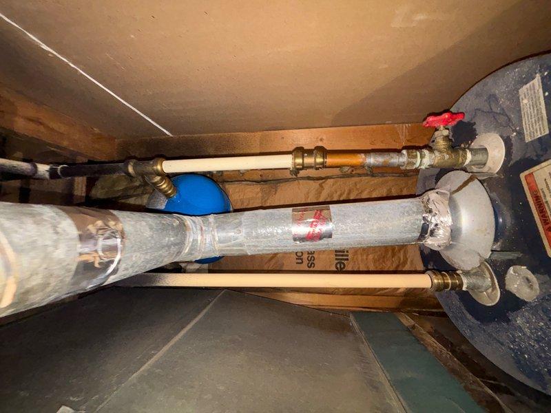 Responded to service call for water heater replacement estimate in attic/crawl space location. Upon arrival, accessed confined utility area and assessed existing water heater installation. Documented current plumbing configuration including copper supply lines with brass fittings, red-handled shut-off valve, white PVC venting system with elbow joints and blue connectors, and metal strapping securing the unit. Observed moisture present on vertical PVC piping and noted opening in ceiling material near installation area. Evaluated structural clearances, wooden joist spacing, and access requirements for 40-gallon capacity replacement unit. Photographed existing installation setup, pipe routing, valve locations, and venting configuration to prepare accurate replacement estimate. Provided customer with assessment findings and replacement recommendations for gas water heater system.