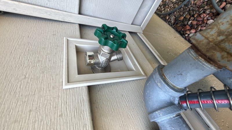 Responded to service call for leaking exterior hose bib. Upon arrival, inspected outdoor water spigot mounted in recessed box on exterior wall and confirmed active leak at valve assembly. Removed damaged brass spigot and assessed supply line condition, noting corrosion on existing vertical pipe. Installed new chrome-plated hose bib with green wheel-style handle in recessed wall box. Connected replacement valve to water supply line and secured all fittings. Tested new installation for proper operation and verified no leaks present. Exterior faucet now functioning correctly with full shutoff capability restored.