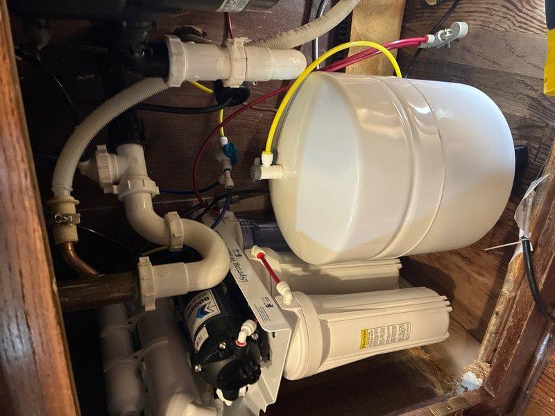 Replaced water filter system under kitchen sink. Removed existing filtration unit and installed new system including white cylindrical storage tank, filtration housing, and associated plumbing connections. Connected supply lines using PVC piping, brass fittings, and flexible hoses with proper clamps and seals. Routed electrical wiring for system operation and verified all connections for proper function and leak-free operation.