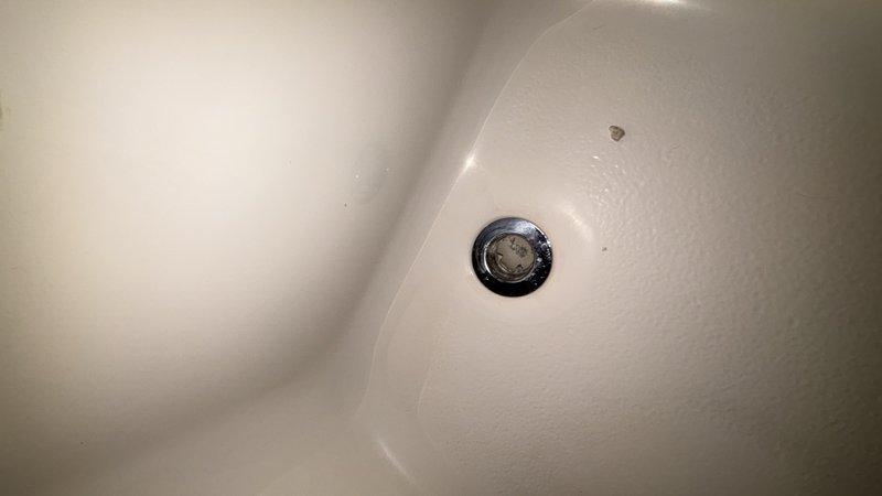 Responded to report of water leaking from upstairs shower through to first floor ceiling. Upon inspection, identified hairline crack in bathtub basin as source of leak. Crack located on interior surface of tub was allowing water to seep through porcelain and penetrate subfloor during shower use. Documented condition of drain assembly and cracked porcelain surface with photographs. Recommended tub replacement or professional refinishing to properly seal crack and prevent continued water intrusion and potential structural damage to ceiling below.