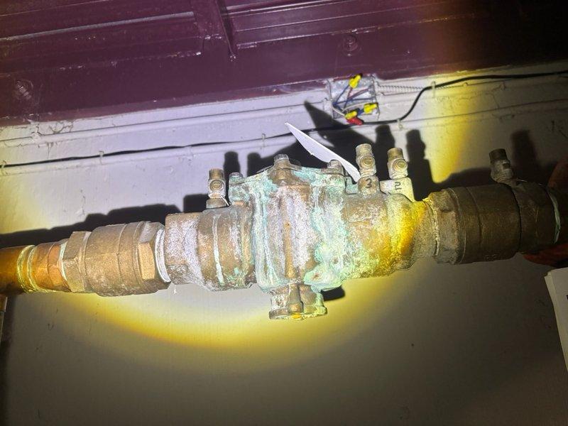 Responded to emergency water leak under stairwell where water was actively spewing from corroded brass piping, requiring building-wide water shutoff. Upon inspection, identified severely corroded brass pipe assembly and pressure reducer valve with extensive verdigris patina and compromised threaded connections. Repaired failed connection point, restored water service to building, and documented existing corrosion on vertical pipe assembly and pressure control components for future maintenance consideration.