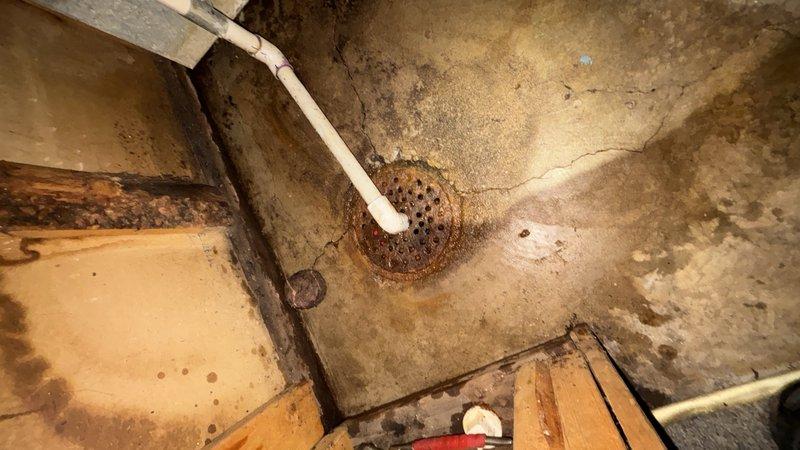 Responded to HVAC recall notice regarding water discharge from floor drain in basement utility area. Upon arrival, inspected floor drain and surrounding concrete surfaces, which exhibited extensive rust-colored staining, deterioration, and water damage radiating outward from drain opening. White PVC drainage pipe observed extending to drain connection point. Concrete floor and walls showed significant moisture exposure, discoloration, and aging consistent with long-term water infiltration. Documented existing conditions including heavily stained drain grate, mottled concrete surfaces with brown and gray discoloration, visible cracks, and worn areas throughout utility space. Area assessed for drainage issues and potential sources of water accumulation at floor drain location.