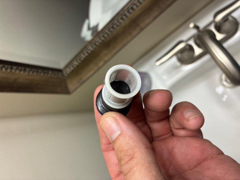 Responded to plumbing maintenance call and performed faucet aerator service at bathroom sink. Removed, inspected, and cleaned clogged aerator assembly which contained debris buildup affecting water flow. The aerator featured a white plastic housing with mesh screening and black rubber gasket that was examined for wear and proper sealing. Component was serviced and reinstalled to restore optimal water pressure and flow pattern at the fixture.