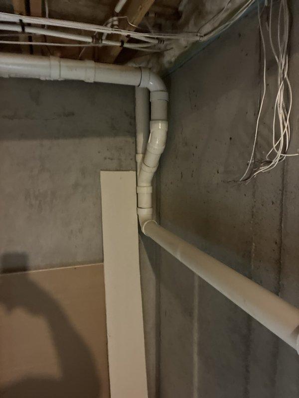 Completed plumbing inspection of basement drainage system, documenting existing white PVC pipe configuration including vertical drain line with multiple elbow joints and horizontal extensions. Photographed pipe installation along concrete walls and verified proper routing of drainage infrastructure in unfinished utility area.