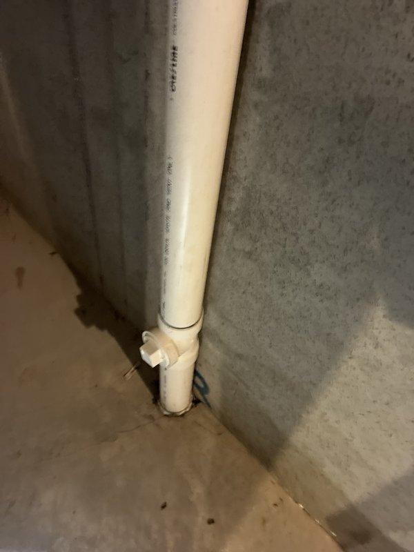 Completed plumbing inspection of basement drainage system, documenting existing white PVC pipe configuration including vertical drain line with multiple elbow joints and horizontal extensions. Photographed pipe installation along concrete walls and verified proper routing of drainage infrastructure in unfinished utility area.