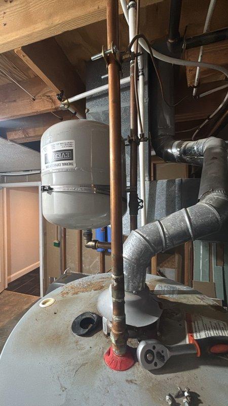 Responded to service call for reported water heater leak at base of unit in basement. Upon arrival, conducted thorough inspection of tanked water heater system and associated plumbing components. Examined white cylindrical water heater tank, expansion tank, copper piping connections, and all valves for signs of active leakage. Observed significant rust staining and discoloration on top crown surface of water heater tank, indicating previous moisture exposure. Inspected base of unit, copper supply lines, pressure relief valve, drain valve, and all threaded connections for source of reported leak. Checked expansion tank mounting and connections. Evaluated condition of oxidized copper piping throughout system. Documented existing rust damage and wear patterns on tank surfaces. Photographed equipment condition and plumbing configuration for service records. Provided assessment of unit condition and discussed repair options with customer to address leaking issue and prevent further water da