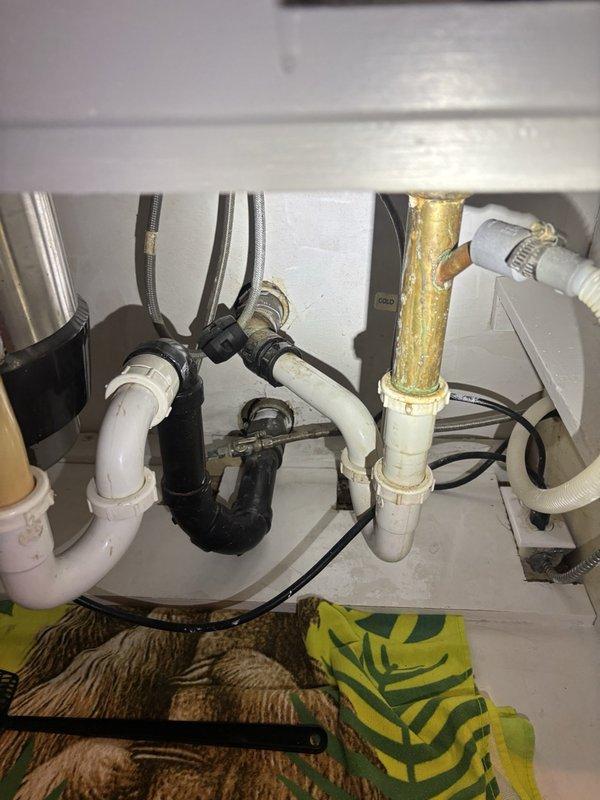 Responded to a clogged kitchen drain line service call. Upon inspection, discovered severely corroded metal drain piping beneath the kitchen sink showing significant rust deterioration and structural compromise. Assessed the existing plumbing configuration, which included a double-basin setup with garbage disposal unit, white PVC drain assemblies, and P-trap connections. Documented the extensive corrosion on the main drain line and confirmed that the existing metal piping has deteriorated beyond repair. Advised that complete drain line replacement will be necessary to restore proper drainage function and prevent future failures. Photographed the corroded pipe sections in the crawl space area and under-sink plumbing assembly for documentation purposes.