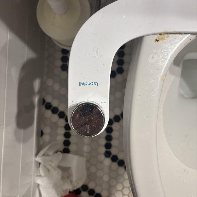 Responded to service call for toilet experiencing repeated clogging issues unrelated to debris or waste material. Upon inspection, found toilet equipped with Brondell bidet attachment showing signs of normal use and minor staining. Evaluated toilet bowl, flush mechanism, and dual-flush button system for proper operation. Assessed water supply line connections and shut-off valve functionality. Determined potential causes of recurring clogs and verified proper water flow through fixture. Documented existing conditions of bidet attachment, toilet assembly, and surrounding plumbing connections for service records.