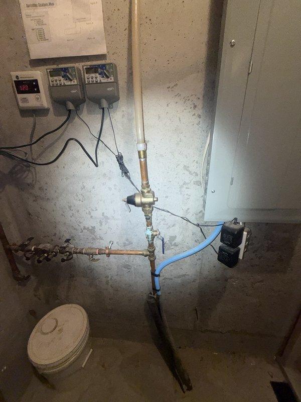 Responded to reported leak originating from above tankless water heater system in basement. Upon inspection, identified the source as plumbing connections associated with the water heater manifold assembly featuring copper and brass piping with T-junction fittings and a blue flexible supply line. Examined all visible pipe connections, valves, and fittings in the vicinity of the digital control units and water heater system. Performed leak detection assessment of the piping assembly to determine exact location and cause of water intrusion, inspecting both the horizontal and vertical pipe runs along with associated shut-off valves and connection points.