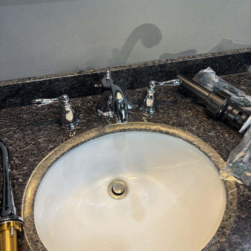 Completed bathroom faucet replacement, installing a new chrome three-piece faucet set with central spout and dual lever handles on granite countertop with undermount sink. Connected new braided stainless steel supply lines with brass compression fittings to shutoff valves, ensuring proper water flow and leak-free operation.