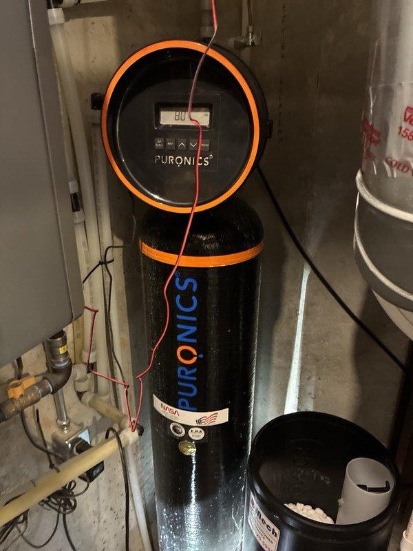 Performed routine maintenance inspection on Puronics water softener system installed in basement. Unit displayed proper operational readings with digital control panel showing normal parameters (80-140 range observed). Verified all electrical connections, including red and blue sensor wiring and power supply cord, were secure and functioning correctly. Inspected plumbing integration points, including copper and plastic pipe connections, valves, and fittings on inlet/outlet lines. Confirmed system components, including orange-banded cylindrical treatment tank and control head assembly, showed no signs of leaks, corrosion, or mechanical issues. System operating as designed with no adjustments or repairs required.