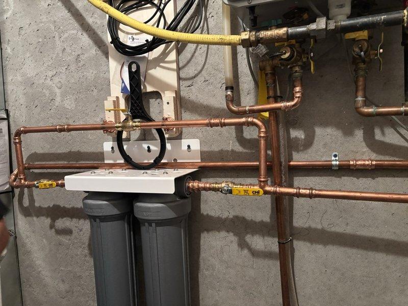 Completed installation of a 2-stage whole home water filtration system with full bypass configuration. Installed dual filter housings with mounting manifold, integrated copper piping with appropriate fittings and valves, and connected electronic control unit for system operation. All components properly secured to wall, bypass valves installed for maintenance accessibility, and system tested for proper flow and function.