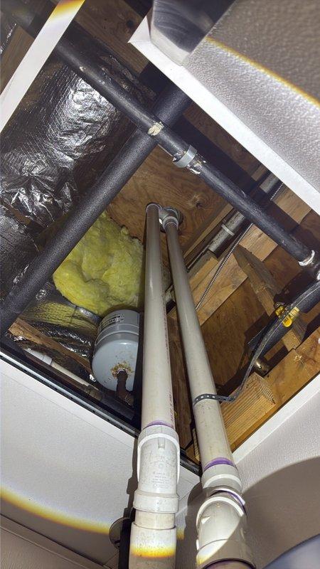 Completed commercial tankless water heater installation featuring a Rinnai unit with full venting and expansion tank integration. Installed white PVC vent piping with proper purple primer application at all joints, routed vertically through ceiling cavity with secure mounting to wooden framing members. Mounted expansion tank to structural joists with appropriate piping connections, coordinated installation around existing HVAC ductwork and insulation materials in confined utility space.