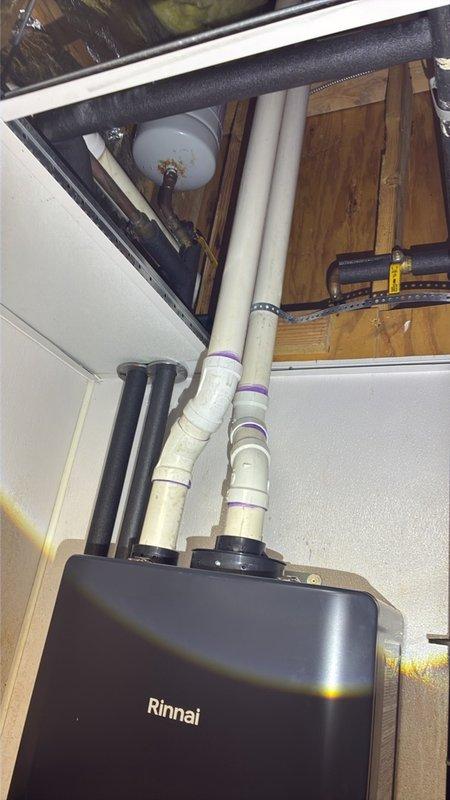 Completed commercial tankless water heater installation featuring a Rinnai unit with full venting and expansion tank integration. Installed white PVC vent piping with proper purple primer application at all joints, routed vertically through ceiling cavity with secure mounting to wooden framing members. Mounted expansion tank to structural joists with appropriate piping connections, coordinated installation around existing HVAC ductwork and insulation materials in confined utility space.