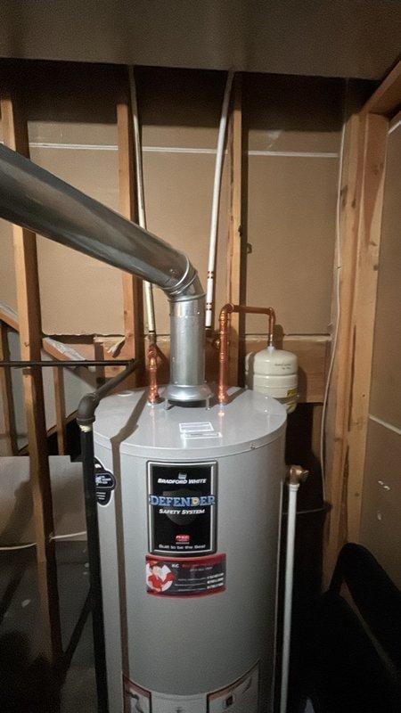 Performed routine plumbing maintenance inspection on DEFENDER Energy System water heater installation. Documented current system conditions including proper venting configuration with metallic exhaust pipe, copper supply line connections, pressure relief valve piping, and white expansion tank mounted on inlet piping. Verified and recorded pressure gauge readings on ProLine gauge showing system operating within normal parameters.