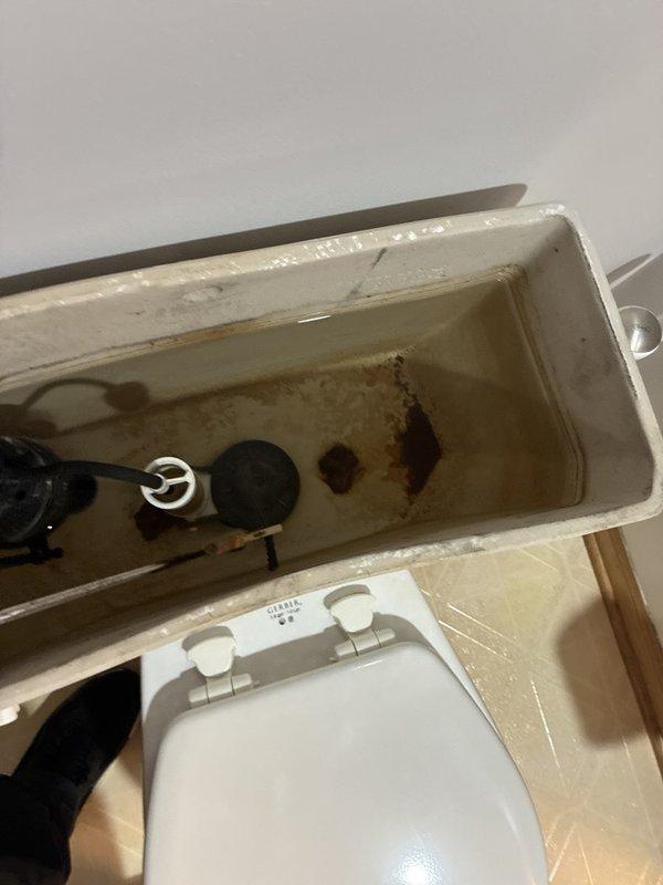 Performed plumbing inspection and documentation of bathroom toilet system. Upon examination, identified significant rust staining and mineral deposit accumulation inside toilet tank, indicating potential internal component deterioration and water quality issues affecting fixture longevity. Assessed flush mechanism and fill valve assembly for operational integrity. Photographed existing conditions for maintenance records, including tank interior deterioration and surrounding wall discoloration near fixture installation point.