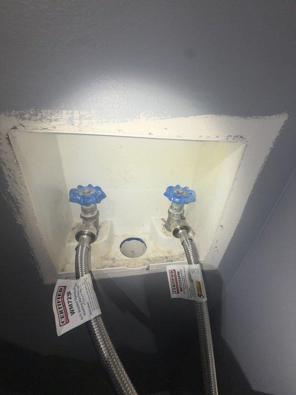 Replaced existing washer box with customer-supplied unit, installing new recessed outlet box with dual shut-off valves featuring blue cross-handle knobs. Connected braided stainless steel flexible supply lines to valves and verified proper drainage connection through center outlet, ensuring secure mounting and leak-free operation for residential washing machine hookup.