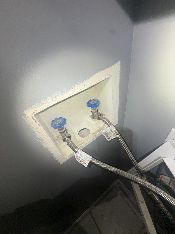 Replaced existing washer box with customer-supplied unit, installing new recessed outlet box with dual shut-off valves featuring blue cross-handle knobs. Connected braided stainless steel flexible supply lines to valves and verified proper drainage connection through center outlet, ensuring secure mounting and leak-free operation for residential washing machine hookup.