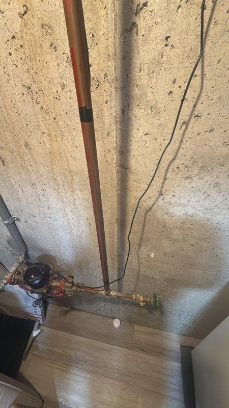 Assessed aging main water shutoff valve assembly in basement for replacement estimate. The existing brass gate valve with red handle shows significant age-related deterioration and tarnishing, requiring modernization to current plumbing standards. Documented existing copper supply line configuration, PVC drainage system, and valve mounting location on concrete foundation wall for preparation of replacement proposal with contemporary quarter-turn ball valve assembly.