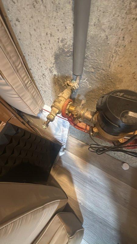 Assessed aging main water shutoff valve assembly in basement for replacement estimate. The existing brass gate valve with red handle shows significant age-related deterioration and tarnishing, requiring modernization to current plumbing standards. Documented existing copper supply line configuration, PVC drainage system, and valve mounting location on concrete foundation wall for preparation of replacement proposal with contemporary quarter-turn ball valve assembly.