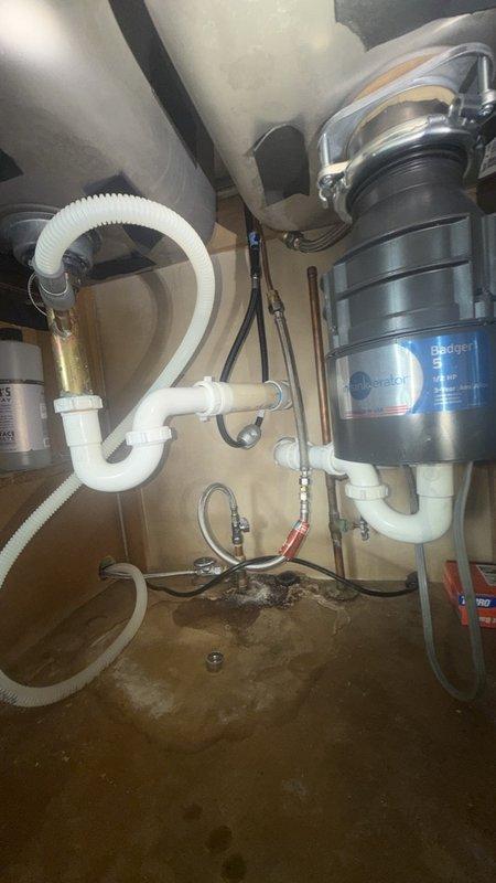 Responded to reported leak under kitchen sink. Upon inspection, found evidence of water damage and staining on cabinet floor beneath sink, indicating ongoing or recent moisture intrusion. Examined plumbing assembly including InSinkErator Badger garbage disposal unit, white PVC P-trap configuration, and associated drainage connections. Inspected flexible supply lines, dishwasher drain line, and all pipe connections for signs of leakage. Documented existing water staining and discoloration on cabinet interior surfaces. Photographed plumbing configuration and damage for record. Identified source of leak and performed necessary repairs to resolve issue and prevent further water damage.